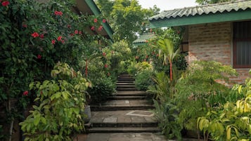 Una escalera de piedra que sube hacia un edificio con techo verde, rodeado de abundante vegetación y plantas con flores.