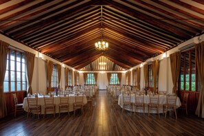 A large hall with high ceilings and wooden beams, arranged with tables and chairs for dining.