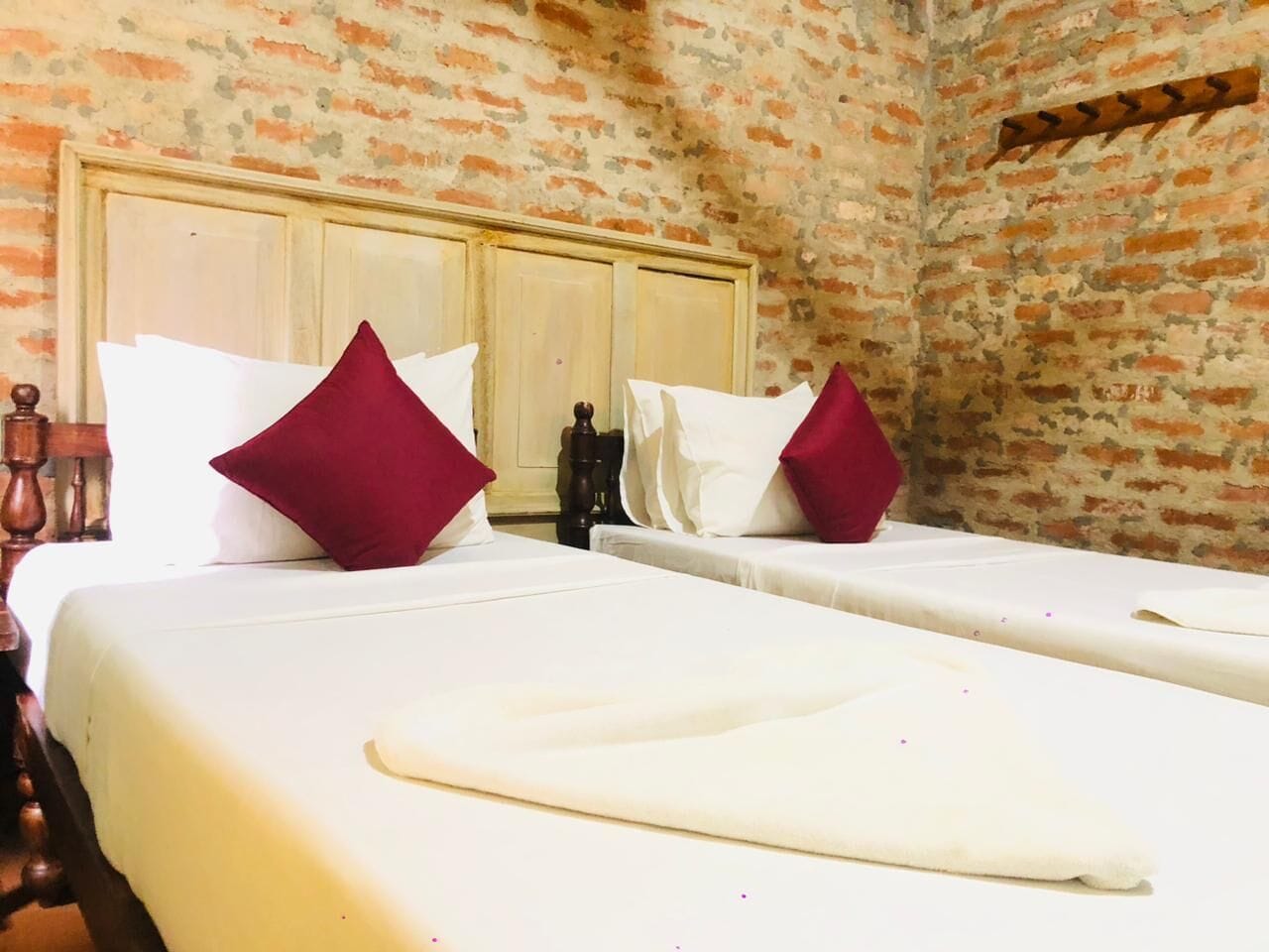 Cama doble con ropa de cama blanca y dos almohadas rojas, cabecera de madera y pared de ladrillo.