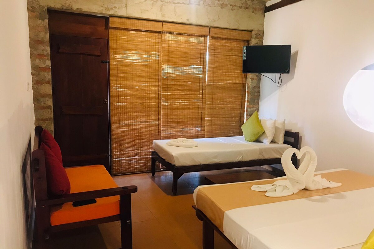 Quarto de hotel com duas camas, televisão presa à parede e porta de correr com persianas de bambu.