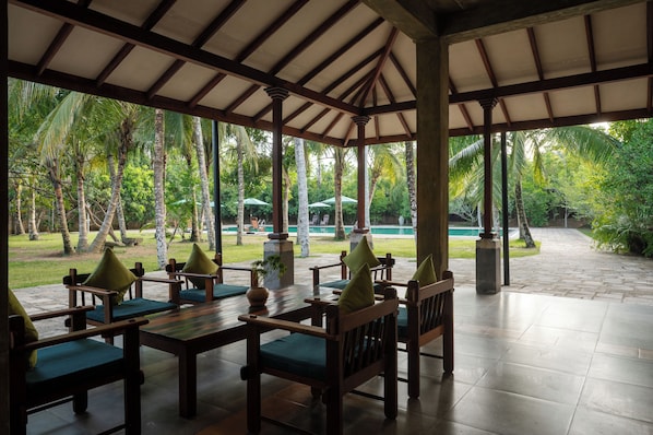 A covered outdoor seating area with wooden furniture and a view of a pool and greenery.