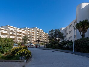 Un immeuble résidentiel à plusieurs étages, doté de balcons, d’un espace de stationnement et d’un jardin bien entretenu agrémenté de fleurs jaunes.