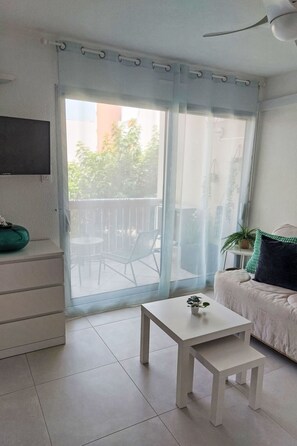 Een kamer met een schuifdeur naar buiten, een witte tafel met een plantje, een bank en een flatscreen televisie aan de muur.