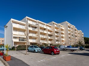 Een meerlaags woongebouw met balkons, een parkeerplaats met auto's, en een heldere blauwe lucht.