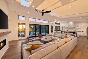 A modern living room with a large sectional sofa, a fireplace, and a dining area with a kitchen in the background. - Lap Pool + Sauna + Outdoor Kitchen | 40 Acres, Glen Ellen | Olive & Oak (Glen Ellen)
