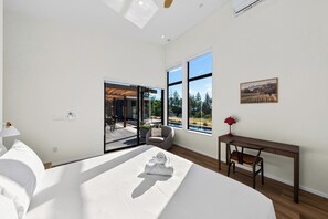 A modern bedroom with a large bed, a desk, and a view of a patio with outdoor furniture.