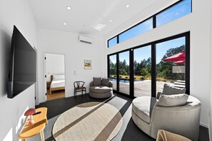 A modern living room with a flat-screen TV, a sofa, a chair, and a large window offering a view of a swimming pool and trees.