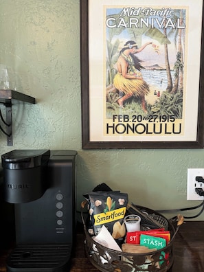 Uma cafeteira Keurig, um cesto com salgadinhos Smartfood e Stash, e um cartaz emoldurado do Carnaval do Pacífico em Honolulu.
