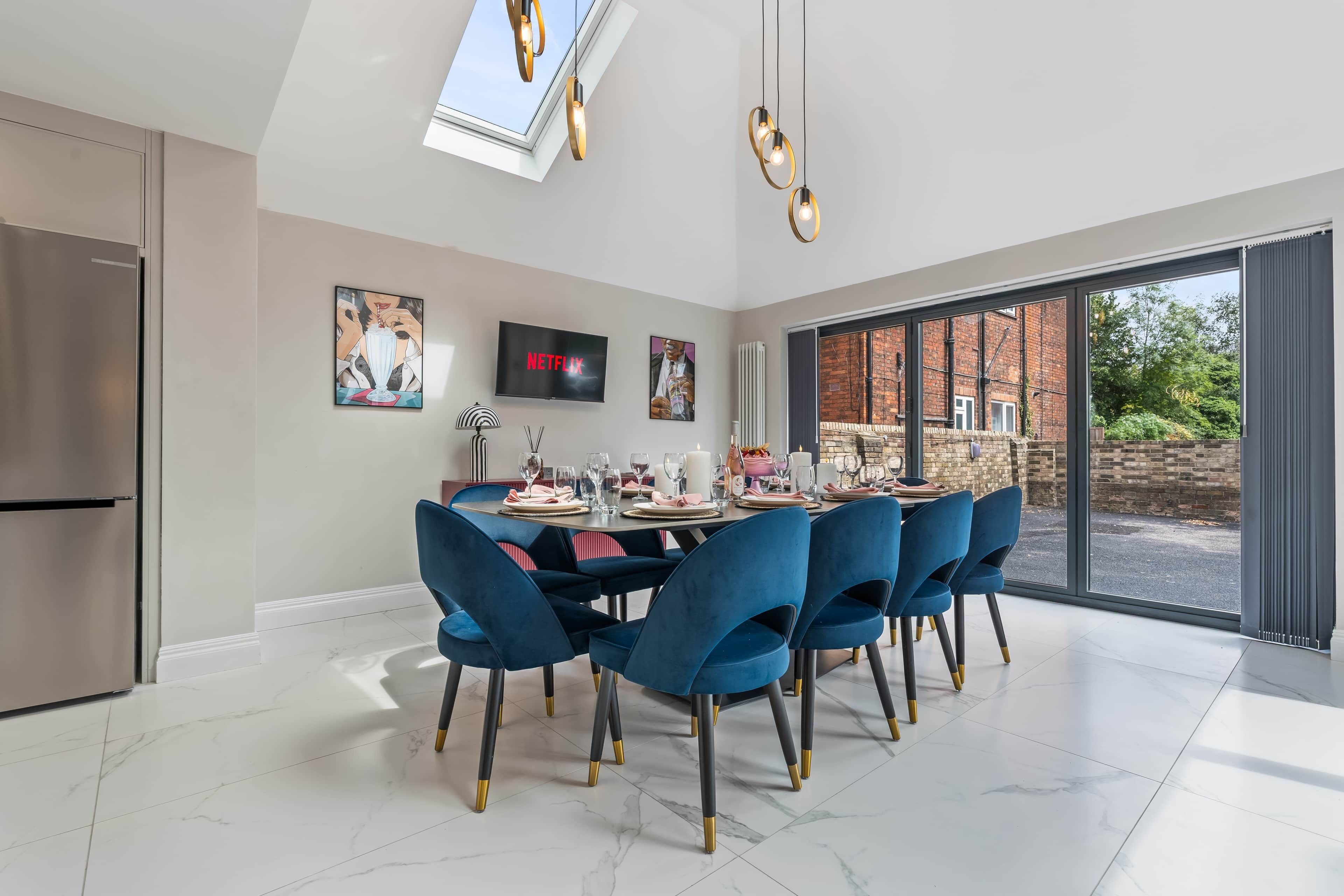 Een moderne eetruimte met een grote tafel, blauwe stoelen, en een televisie waarop 'NETFLIX' wordt weergegeven.