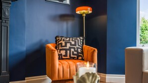 Une pièce avec un mur bleu foncé, un fauteuil orange agrémenté d’un coussin à motifs, un lampadaire et une table sur laquelle se trouvent une bougie et des objets décoratifs.