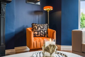 Una habitación con una pared azul oscuro, un sillón naranja con un cojín estampado, una lámpara de pie y una mesa con una vela y adornos.
