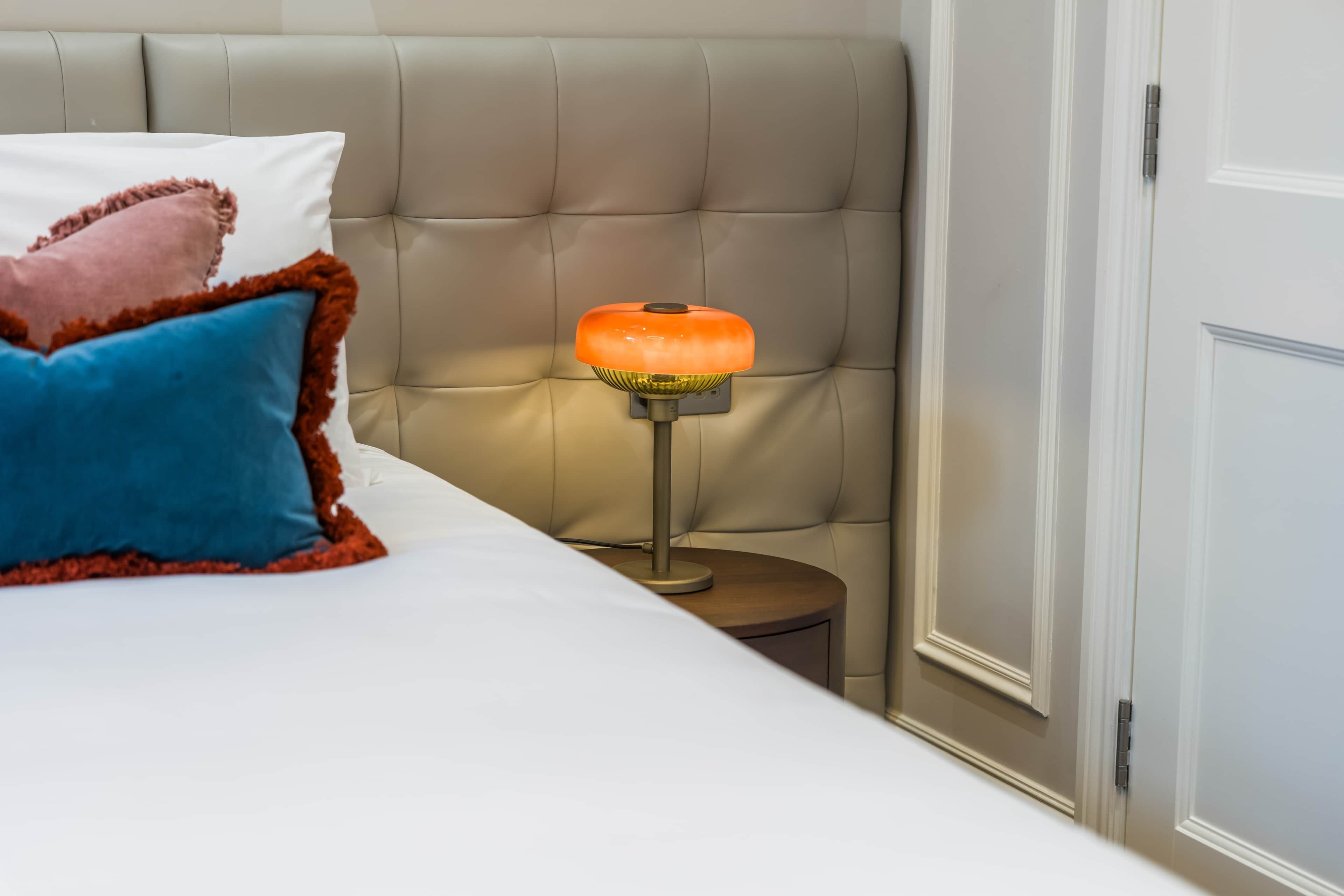 A bed with a white sheet, a blue pillow, and a pink pillow with a brown trim. There is a small wooden nightstand with a lamp that has an orange shade.