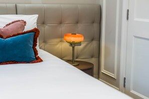 A bed with a white sheet, a blue pillow, and a pink pillow with a brown trim. There is a small wooden nightstand with a lamp that has an orange shade.