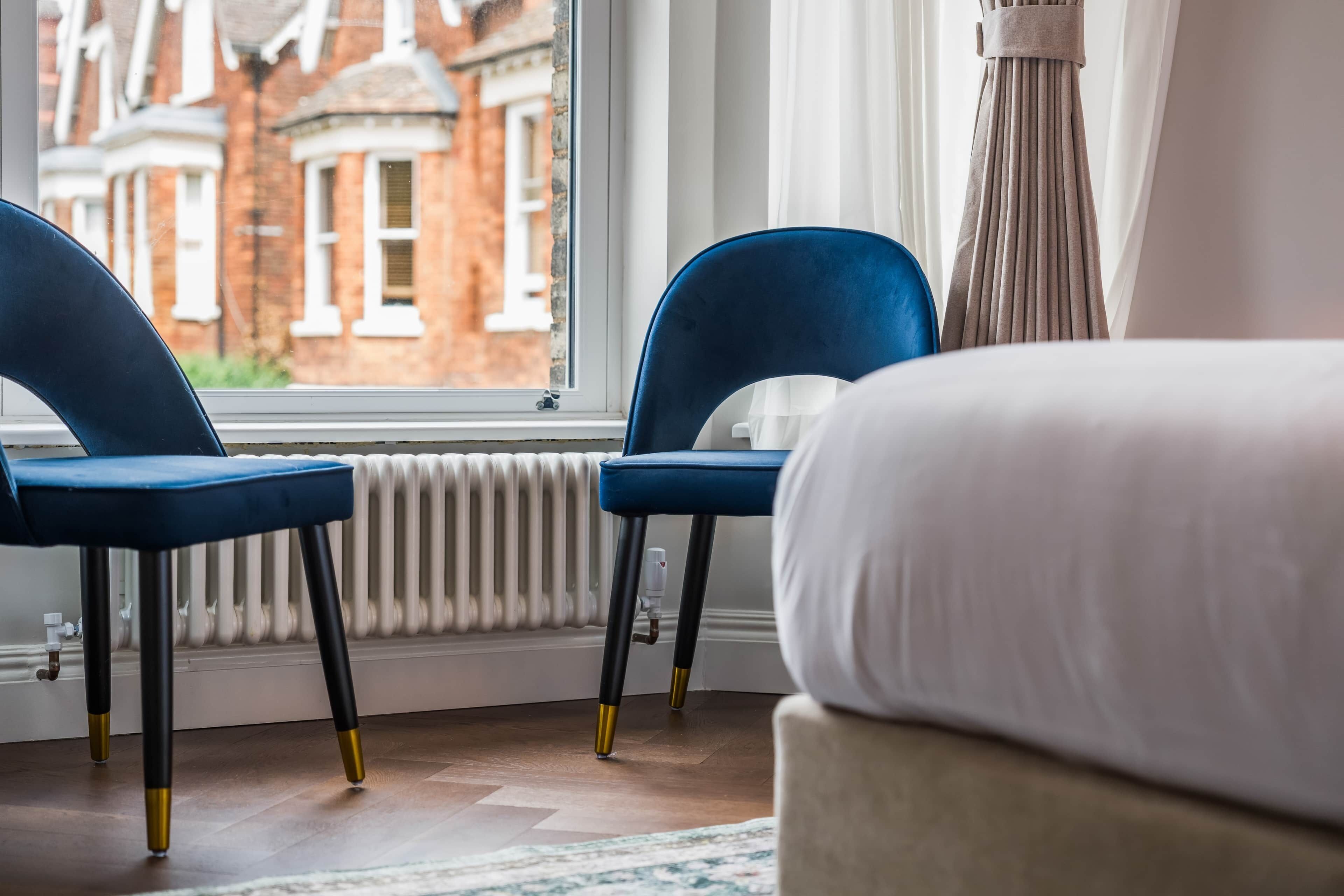 Una stanza con due sedie blu, un letto bianco, un termosifone e vista su un edificio in mattoni rossi attraverso una finestra.