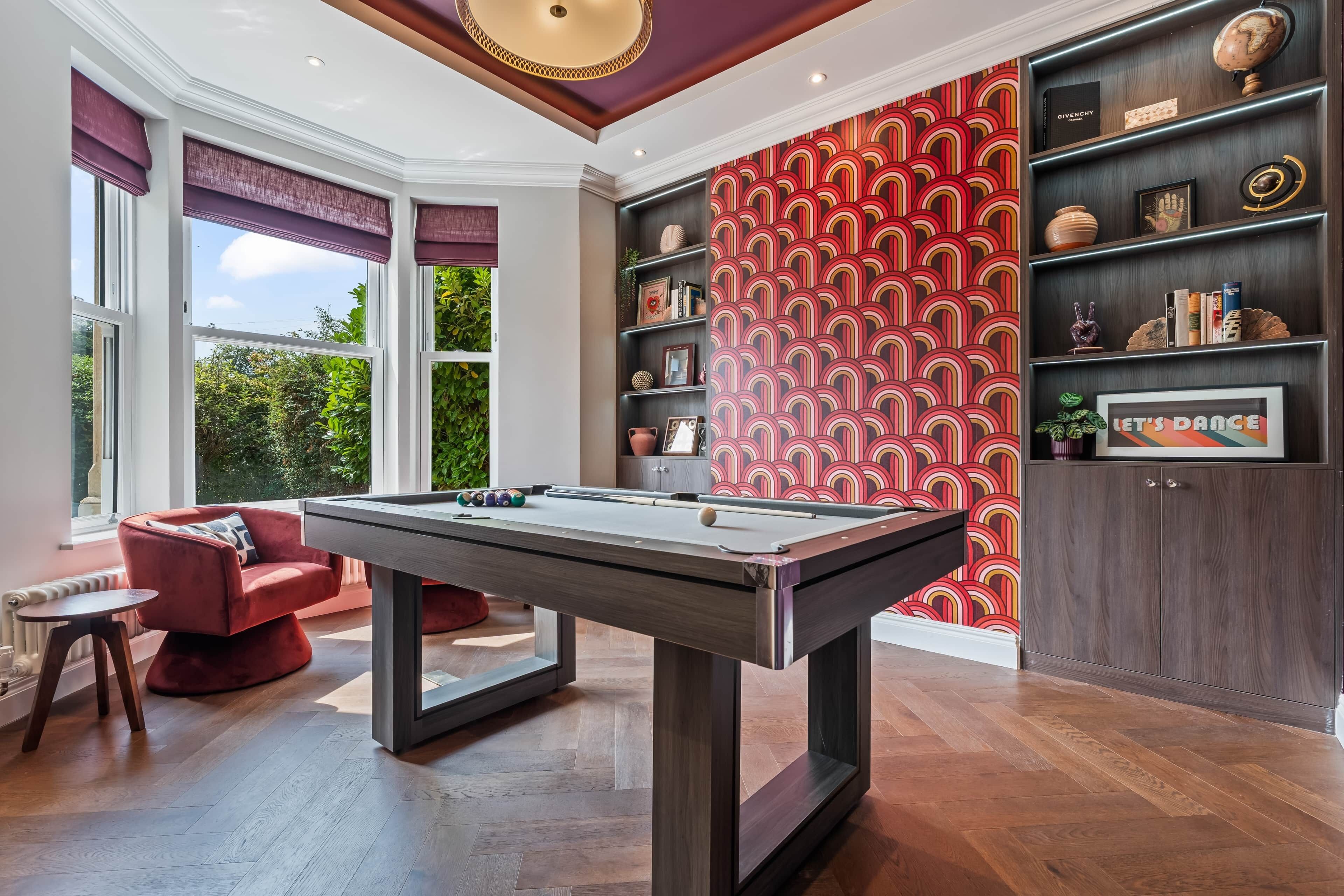 A billiards table in a room with a view of greenery, a red patterned wall, and a bookshelf with various items.