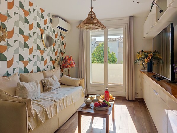 A living room with a sofa, a table with fruits and a bowl, a hanging lamp, and a TV on the wall. - Les Lavandes by Interhome (Saint Cyr sur mer Les Lecques)