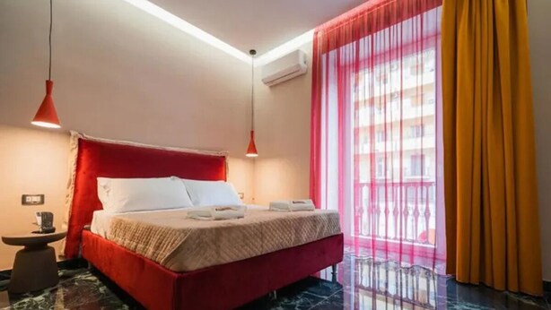 Una camera d'hotel con un letto, un comodino, una testiera rossa, una finestra con tende leggere e una lampada a sospensione rossa.