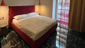 Camera d'albergo con un letto, un comodino, una testiera rossa, una tenda rossa e vista sull'esterno.