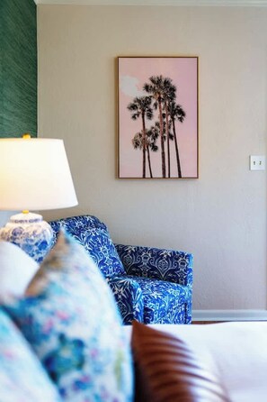 A room with a bed, a blue patterned sofa, a wall-mounted picture of palm trees, and a lamp. - Swank Suites | King with Lux Tub (Ashland)