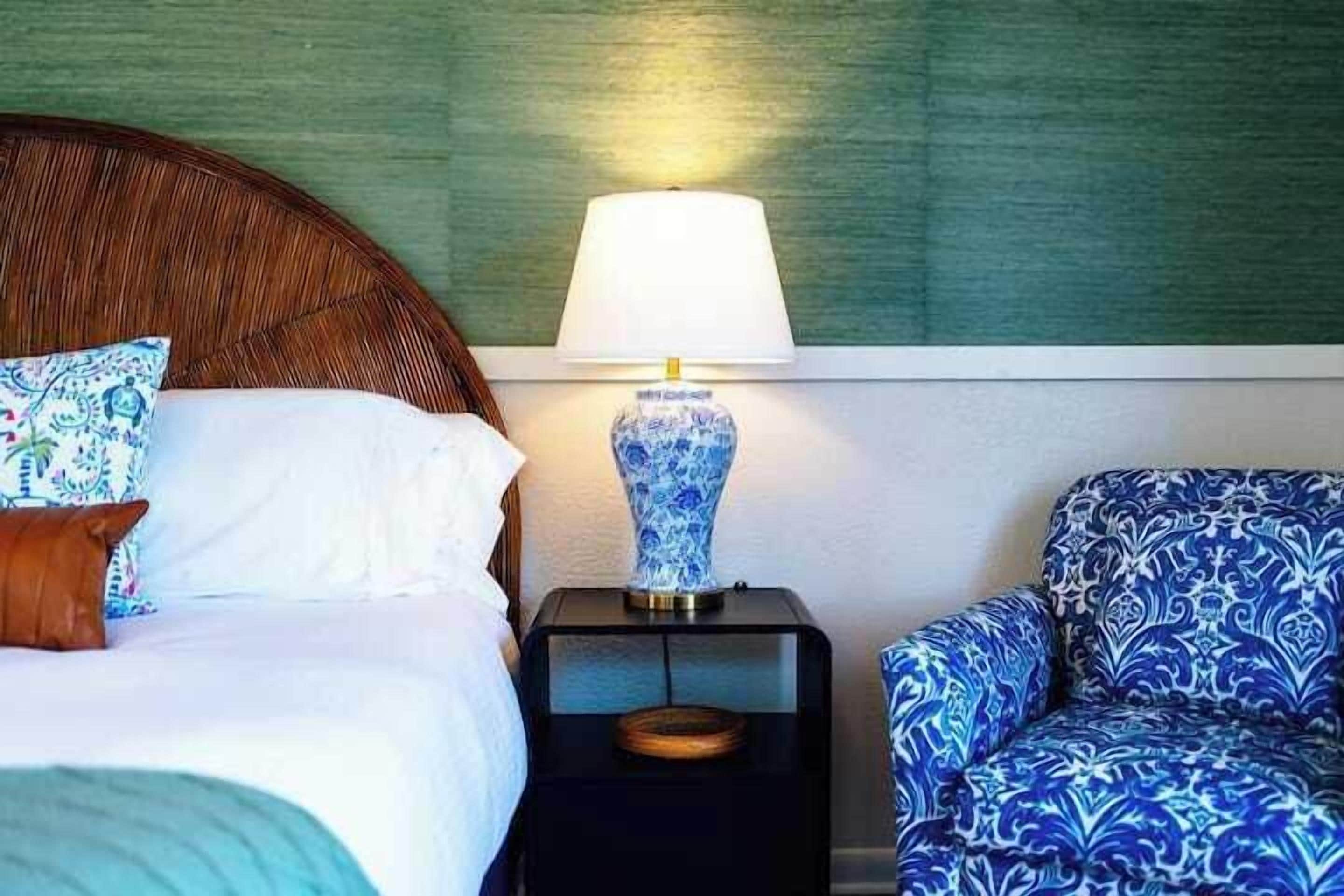 Quarto de hotel com uma cama, mesa de cabeceira com abajur e uma poltrona estampada.