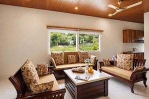A living room with a sofa set, a coffee table, and a window with a view of greenery. - Karinya Villas Scenic Hill View & Garden (Naina Range)