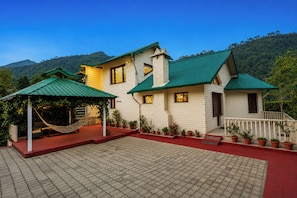 A two-story house with a green roof, a covered patio with a hammock, and potted plants. - Karinya Villas Scenic Hill View & Garden (Naina Range)