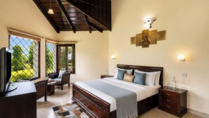 A hotel room with a bed, bedside tables, a desk with a chair, a ceiling fan, and large windows with a view of greenery. - Karinya Villas Scenic Hill View & Garden (Naina Range)
