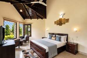 A hotel room with a bed, bedside tables, a desk with a chair, a ceiling fan, and large windows with a view of greenery.