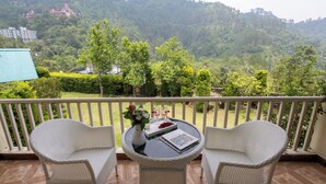 A balcony with wicker chairs, a small table with a book and flowers, and a view of a forested hillside. - Karinya Villas Scenic Hill View & Garden (Naina Range)