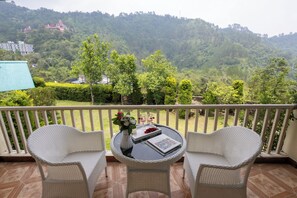 A balcony with wicker chairs, a small table with a book and flowers, and a view of a forested hillside.