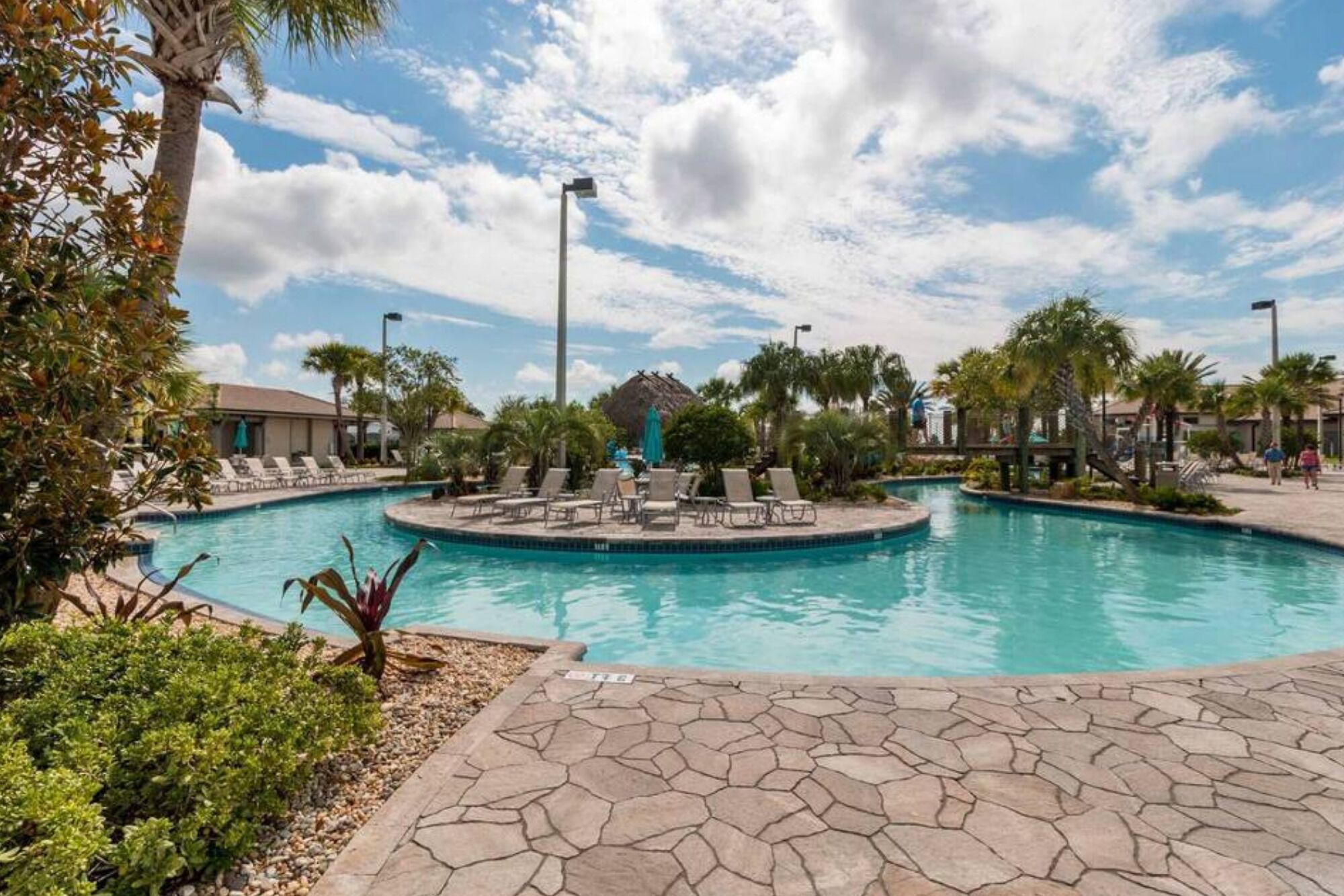 A pool area with lounge chairs and a small building, surrounded by palm trees and a clear sky.