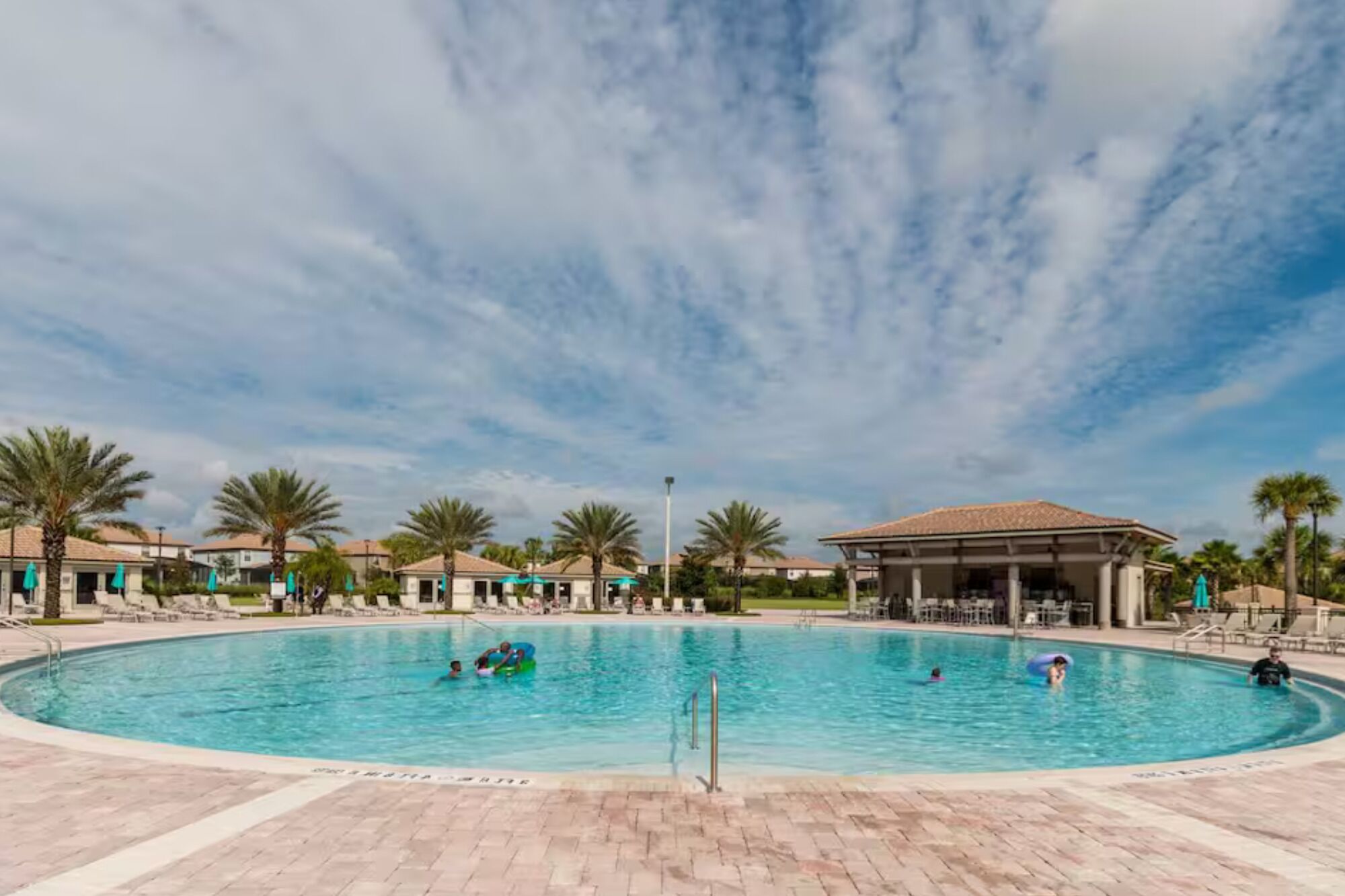 A swimming pool with people in the water, surrounded by palm trees and buildings.