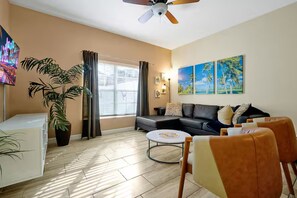 A living room with a sofa, chairs, a coffee table, a TV, a ceiling fan, a window with curtains, and wall art. - Private 4BR Retreat with Pool Near Disney (Championsgate)