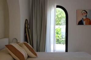 Une chambre à coucher avec un lit, un tableau, une fenêtre avec des rideaux et une vue sur la verdure à l’extérieur.