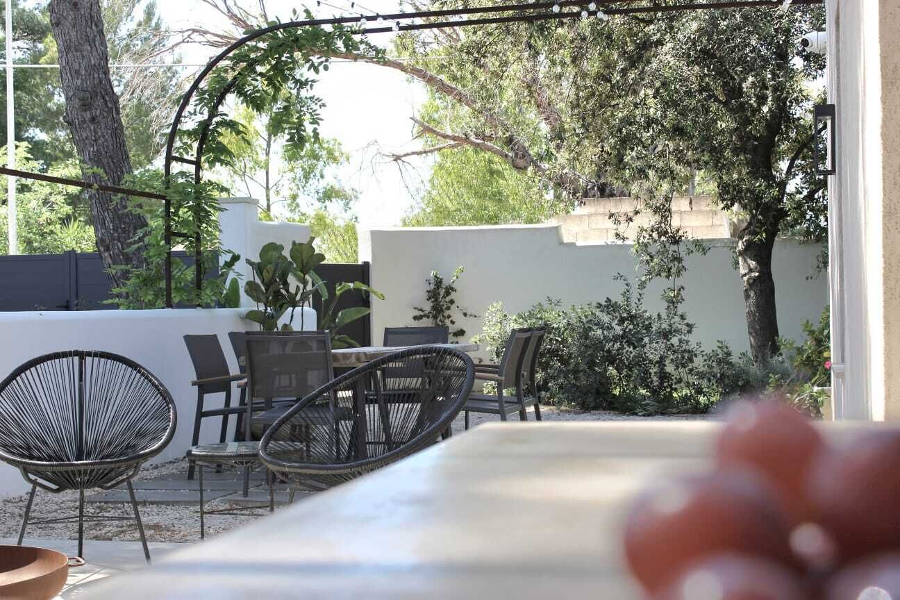 Um pátio externo com mesa e cadeiras, rodeado por vegetação e uma parede branca.