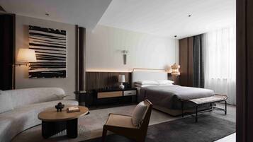 Una habitación de hotel moderna con sofá, una mesita, una silla, una cama, una mesita de noche, una lámpara y una obra de arte en la pared.