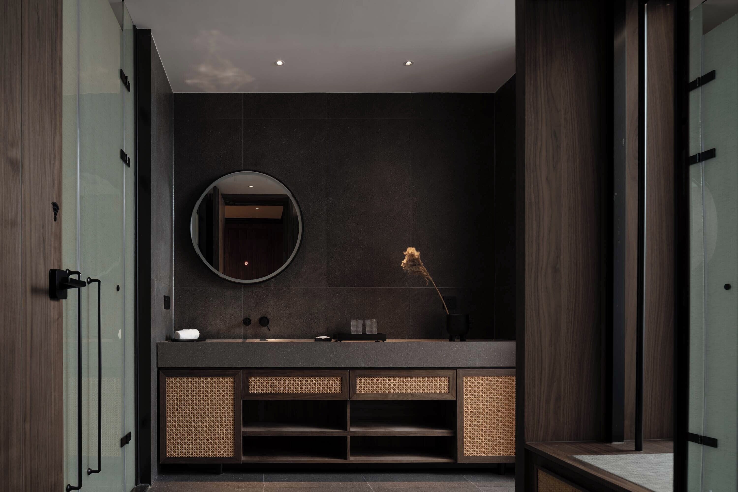 Un baño moderno con una pared de azulejos oscuros, un espejo redondo, un lavabo con encimera oscura y una mopa en el suelo.