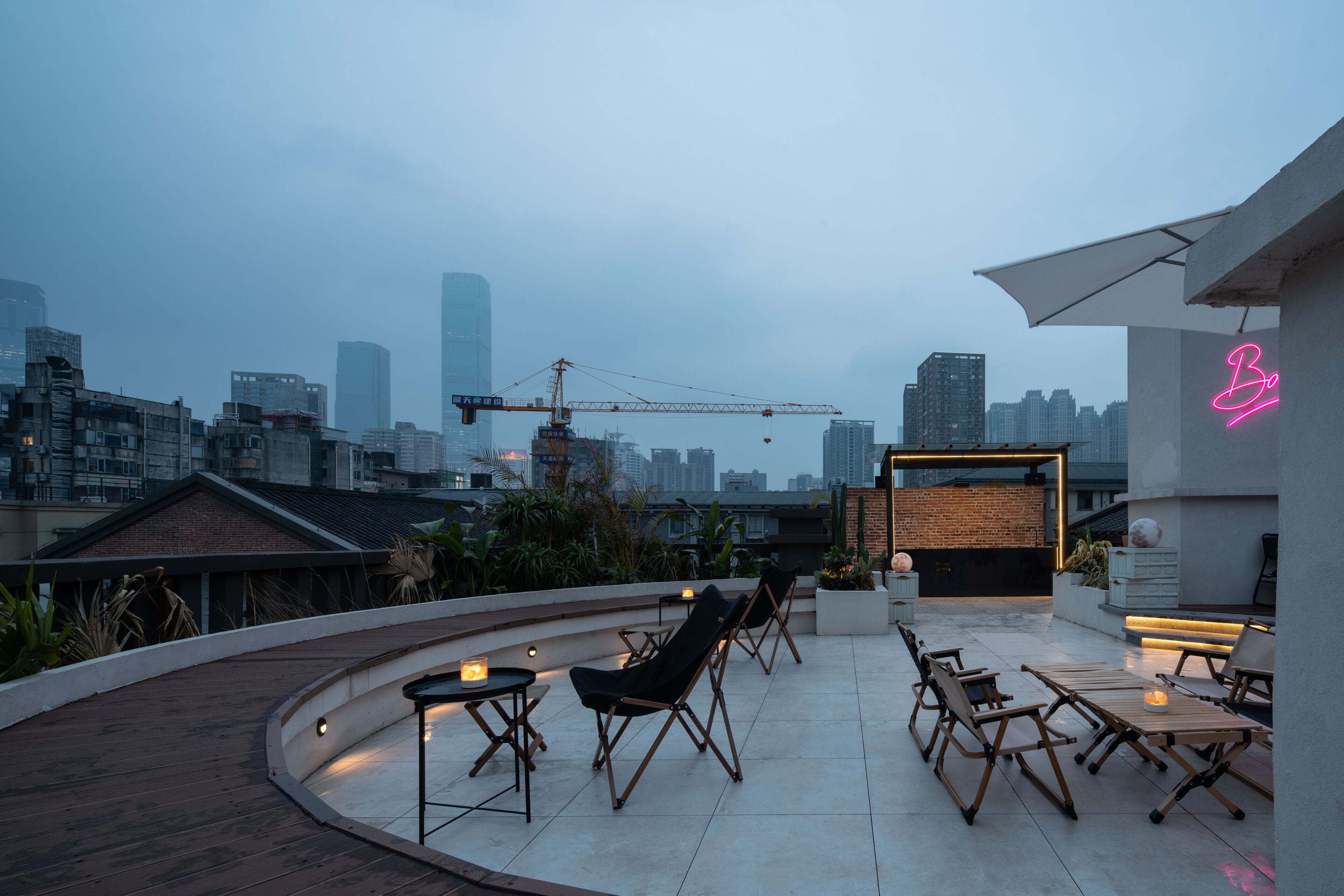 A rooftop terrace with seating and a neon sign, overlooking a cityscape with buildings and construction cranes.