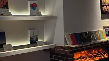 Una habitación con chimenea de ladrillo, estanterías blancas con libros y juegos, y un cuadro en la pared.