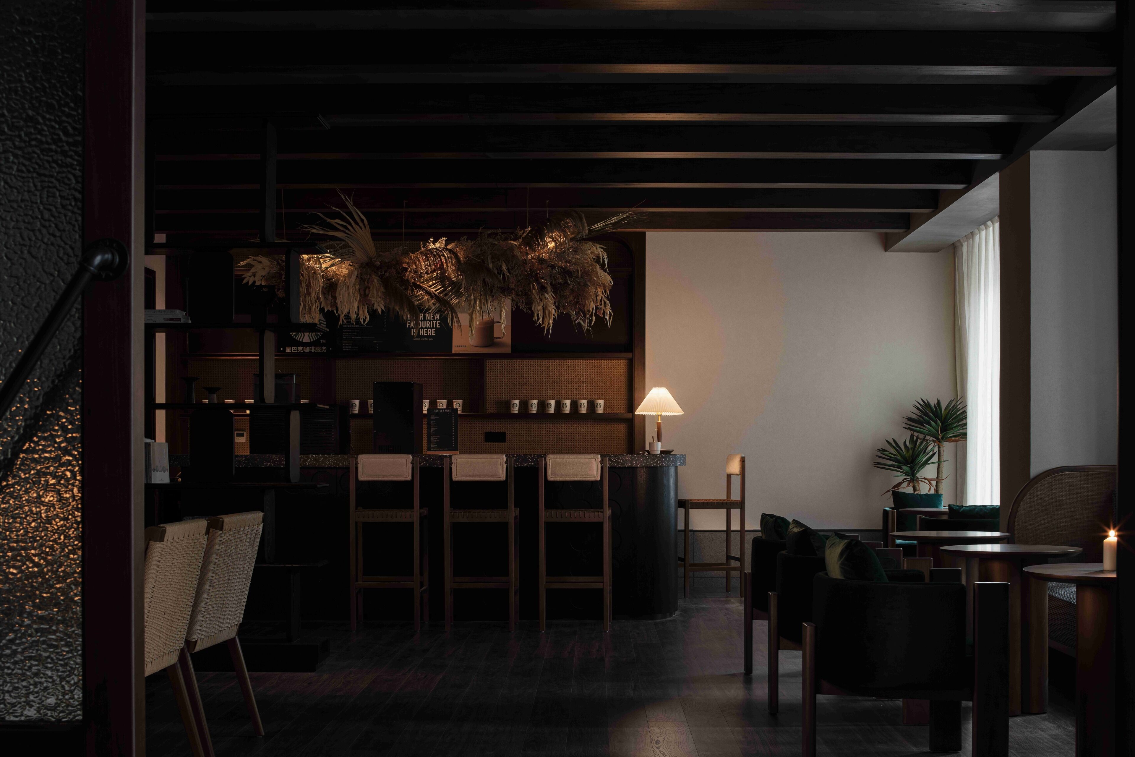 Zona de bar tenuemente iluminada con barra, sillas y una mesa. Hay plantas en macetas y una lámpara en la barra.