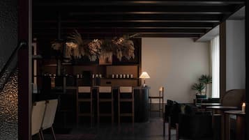 Zona de bar tenuemente iluminada con barra, sillas y una mesa. Hay plantas en macetas y una lámpara en la barra.