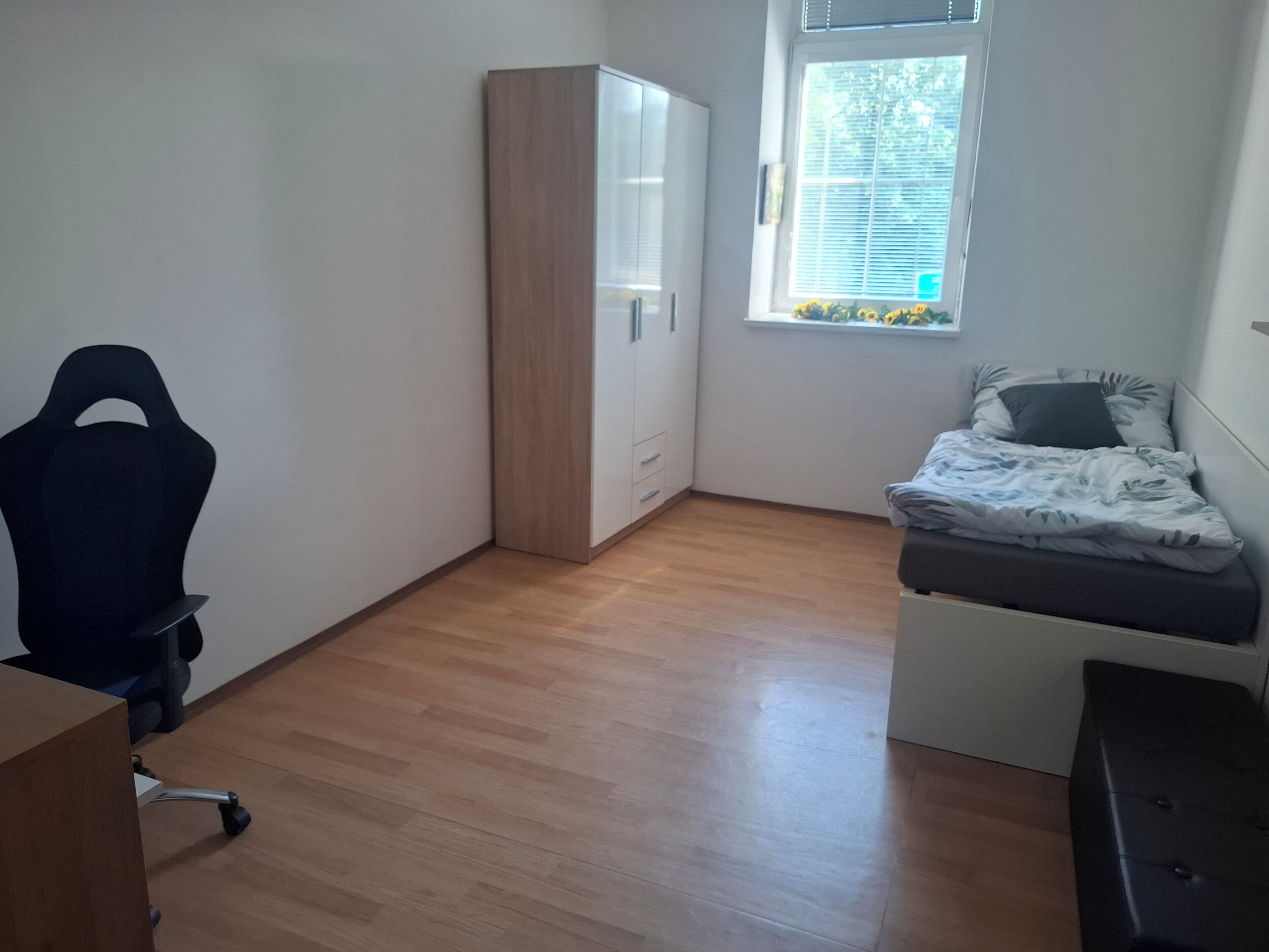 Una habitación pequeña y luminosa, con suelo de madera, una cama con manta estampada, un escritorio con silla y un armario.