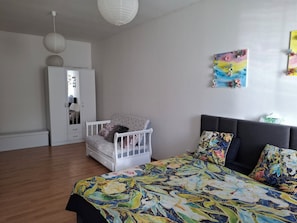A bedroom with a bed, a white chair, a wardrobe, and a wall with a floral painting.