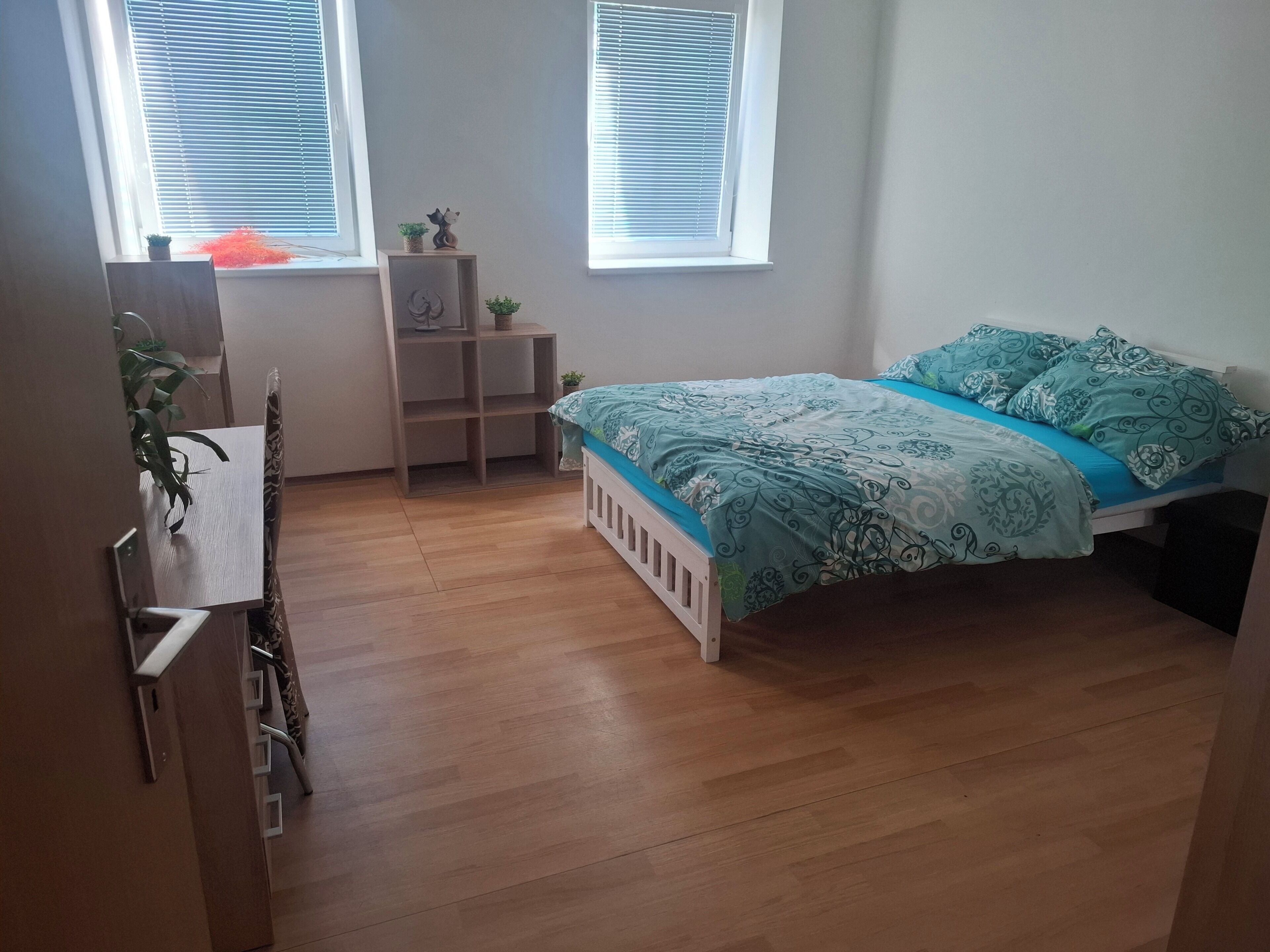 Un dormitorio con cama, escritorio, silla, estantería con plantas y dos ventanas con persianas.