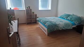 Un dormitorio con cama, escritorio, silla, estantería con plantas y dos ventanas con persianas.