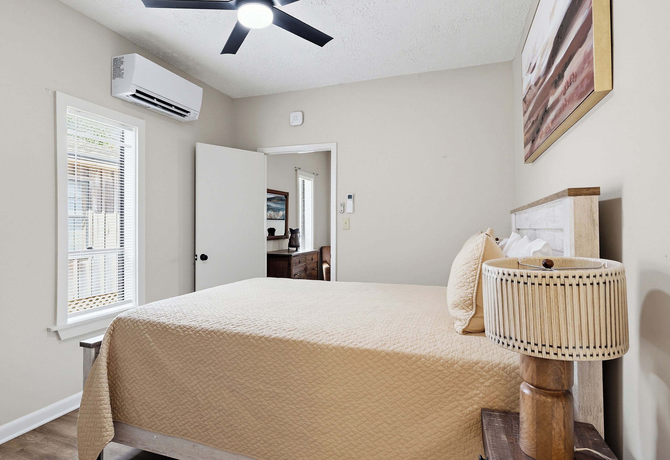 Un dormitorio con cama, ventilador de techo, aire acondicionado, ventana con persianas, lámpara de noche y puerta que da a otra habitación.