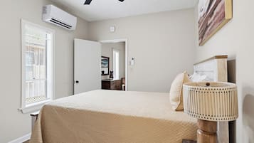Un dormitorio con cama, ventilador de techo, aire acondicionado, ventana con persianas, lámpara de noche y puerta que da a otra habitación.