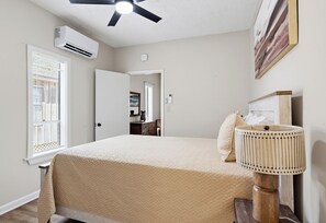 A bedroom with a bed, a ceiling fan, an air conditioning unit, a window with blinds, a bedside lamp, and a door leading to another room.