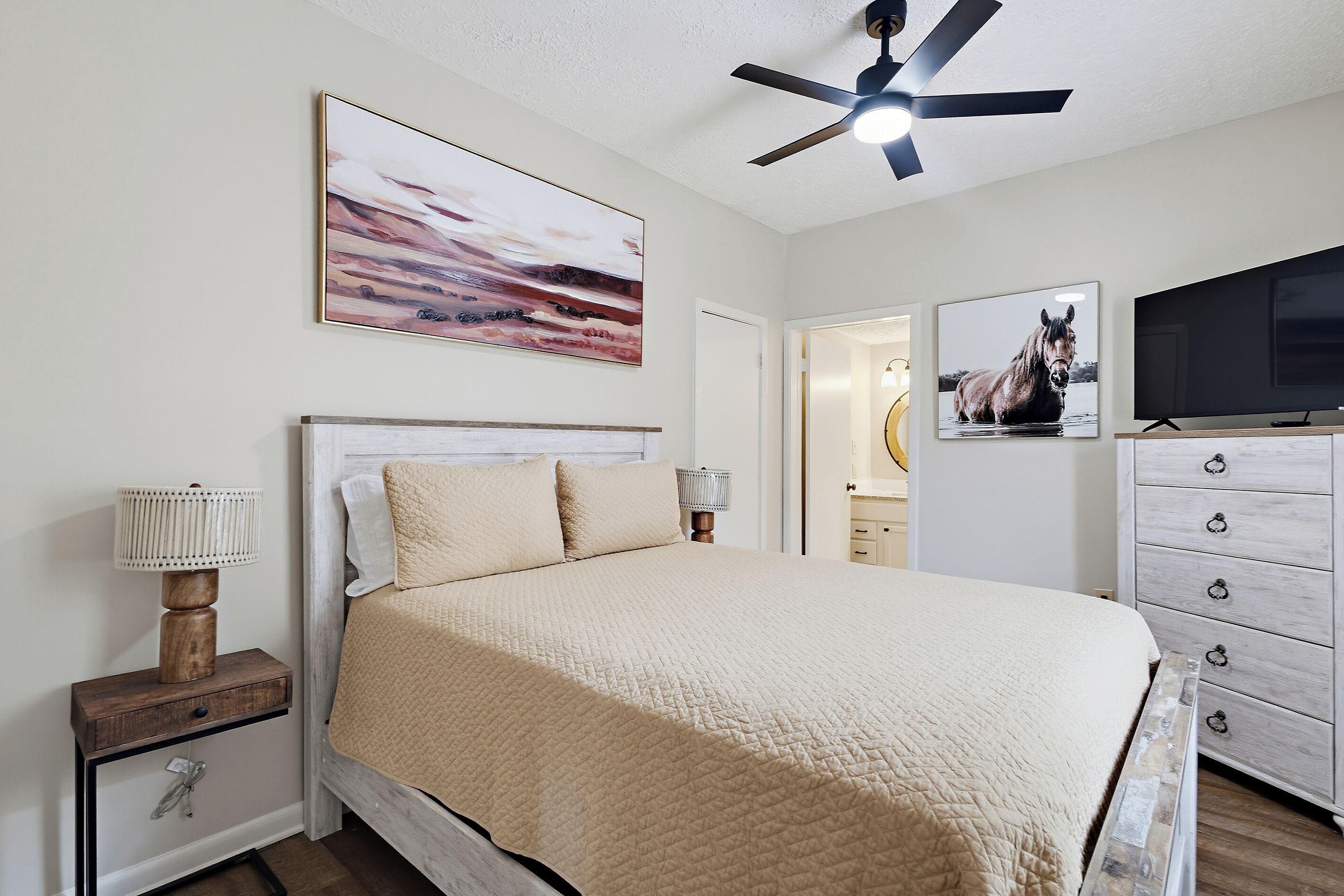 A bedroom with a bed, a ceiling fan, a TV, and artwork on the walls.