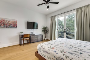 Ein Schlafzimmer mit einem Bett, einem Schreibtisch, einem Stuhl, einer Kommode, einem Fernseher, einer Pflanze und einer Glastür zu einem Balkon.